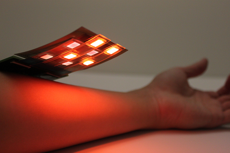 A thin, flexible electronic board, lit-up with red and infrared lights, and approximately three inches wide by three inches wide, is held above a person's forearm.