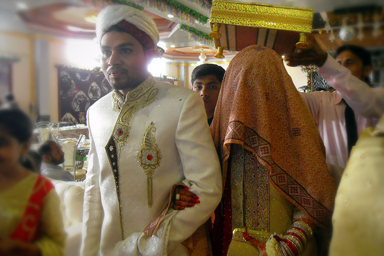 A Pakistani couple in elaborate ceremonial wedding wear. The woman's entire head is covered by a cloth.