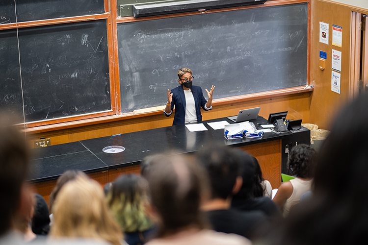 Associate professor Poulomi Saha lectures her large class on cults in popular culture. She is in front of a large blackboard with her hands raised to her shoulders.