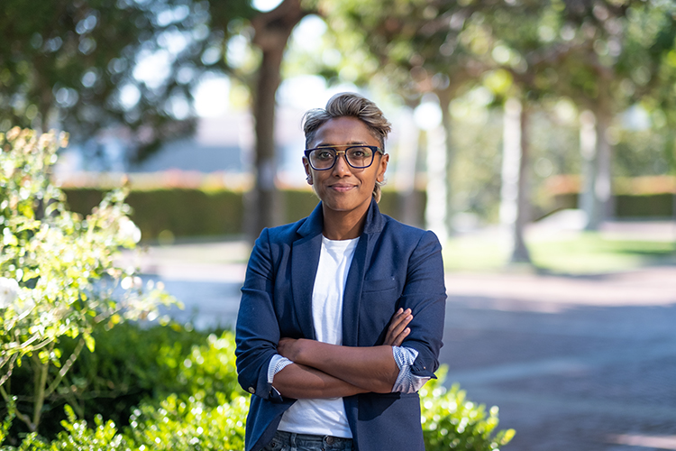 A portrait of associate professor of English Poulomi Saha, who is standing with her arms crossed wearing a white T-shirt and a blue blazer. She has large eyeglasses and a short haircut.