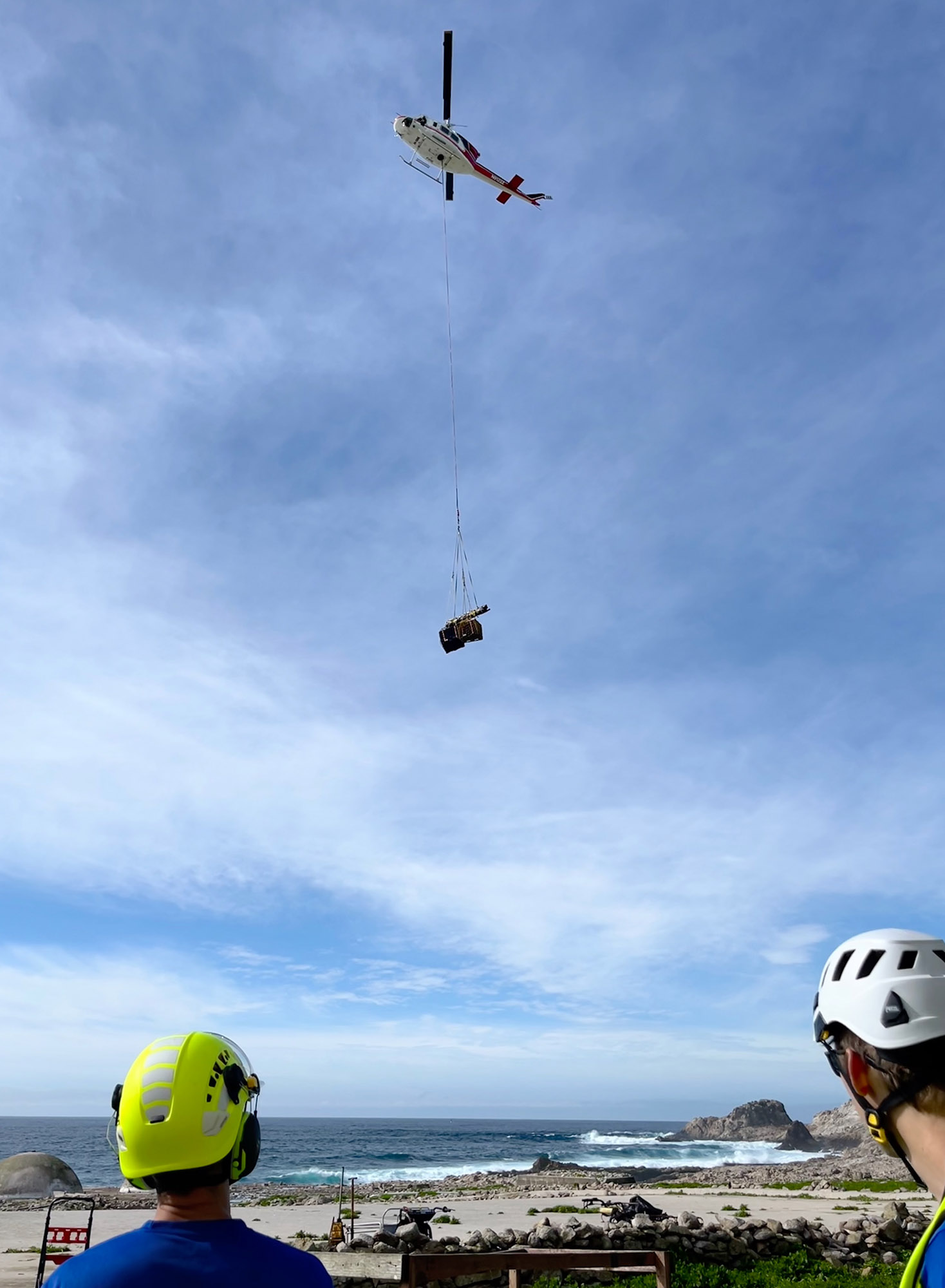 a net containing equipment dangles from a helicopter as two people in hard hats look on from the ground
