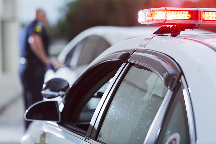 A police officer talks to a driver who he's pulled over for speeding. The lights on his police car are flashing.