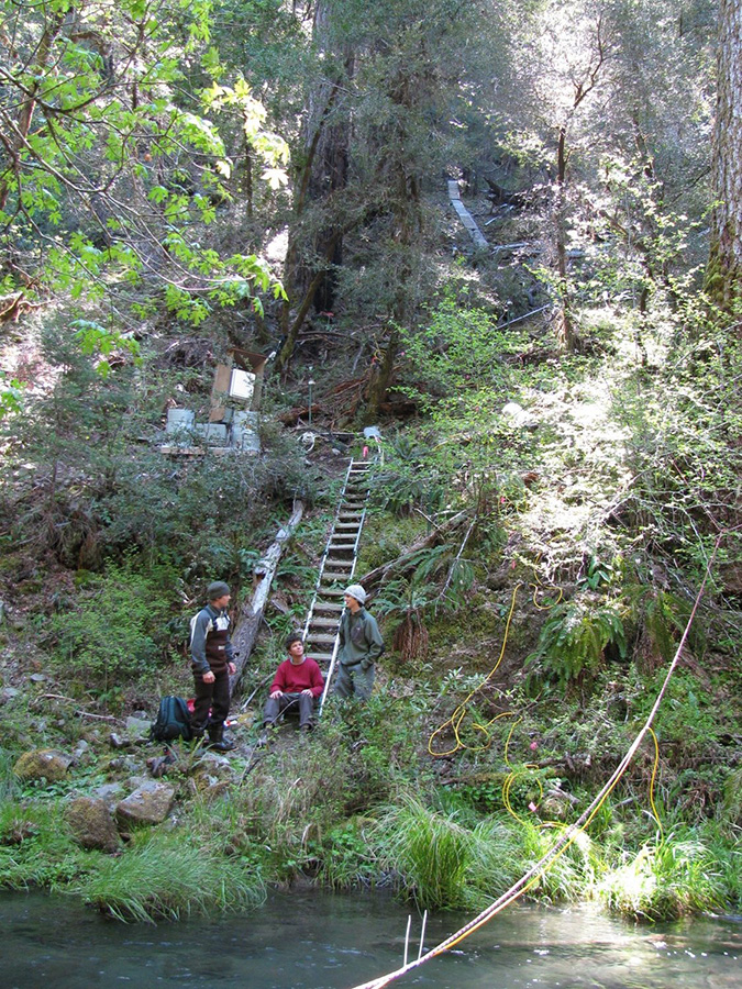 Researchers on a slope next to a river, one is sitting on a ladder