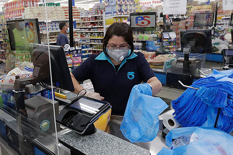 A retail checkout clerk, masked against coronavirus infection, puts purchases into blue plastic bags