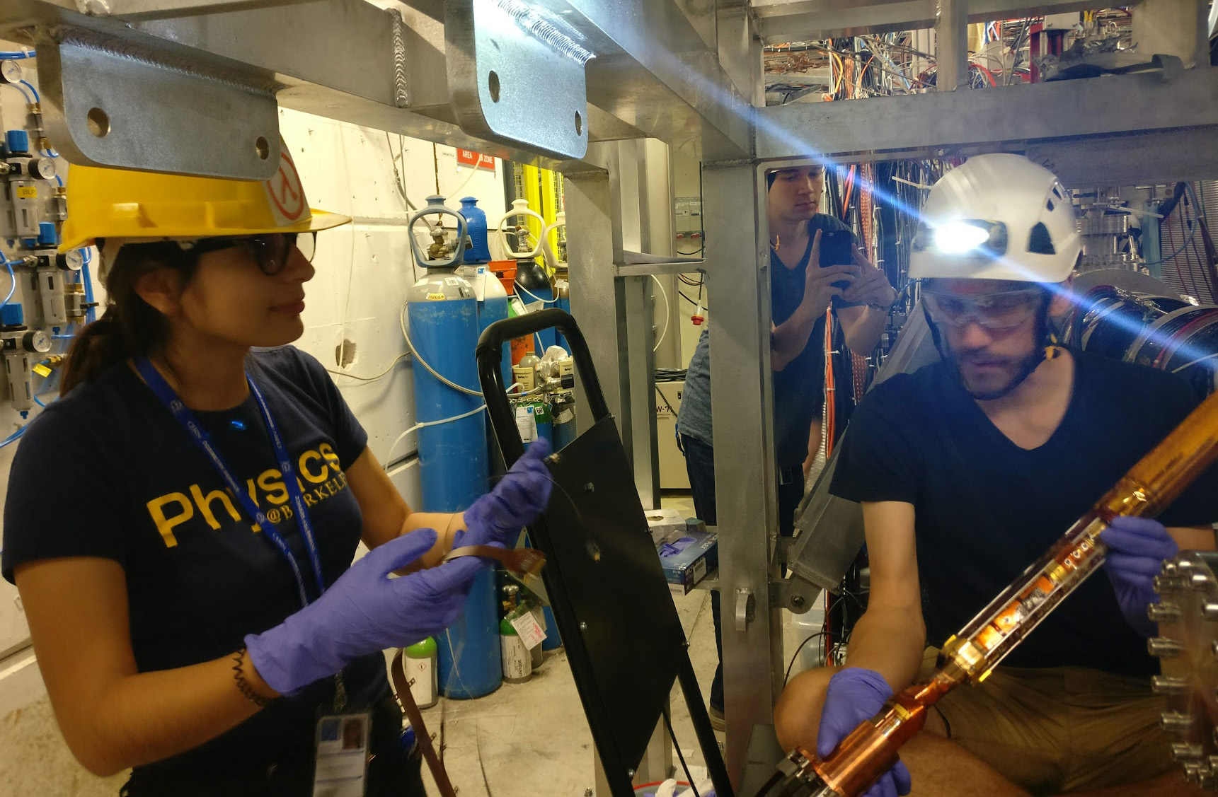 Young woman in yellow hardhat and blue T-shirt with Physics@Berkeley written on it, working beside a bearded man holding a copper-plated tube