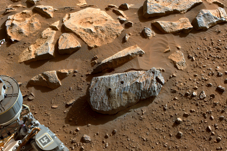 closeup of rock on Mars sitting in red sand with wheel of rover at lower left