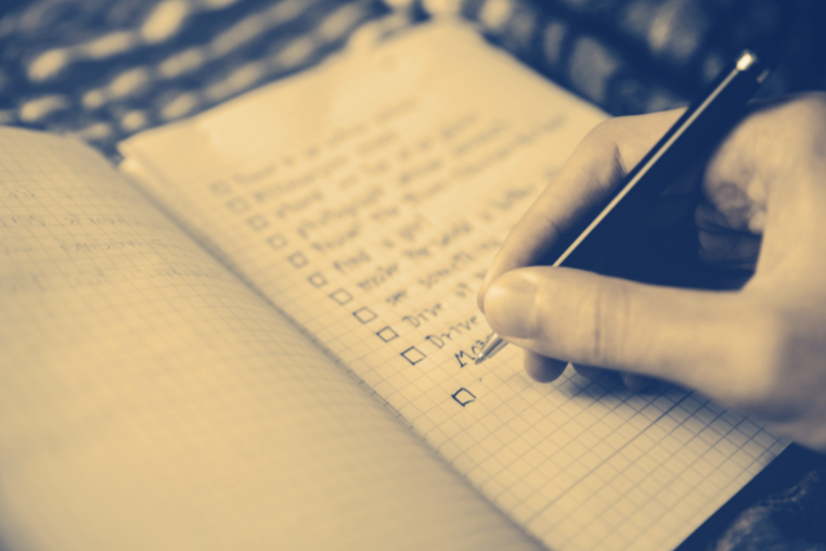 A person's hand writing a list of items on a piece of paper next to boxes that will be checked off.