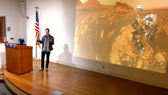 Sergey Levine speaks from a stage with a large screen projecting the Mars rover in the background