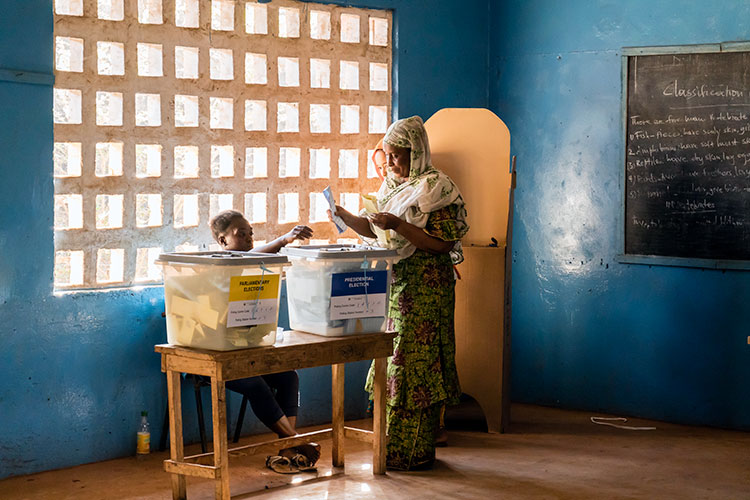 In a blue, sunlit room, a woman casts her vote in the Sierra Leone presidential election