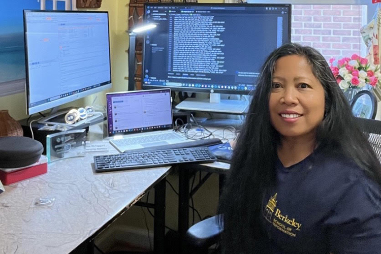 SupraptiMcTaggart, an alumna of UC Berkeley who was trained at the Citizen Clinic, sits at a desk with several computer monitors and smiles at the camera wearing a Berkeley School of Information T-shirt.
