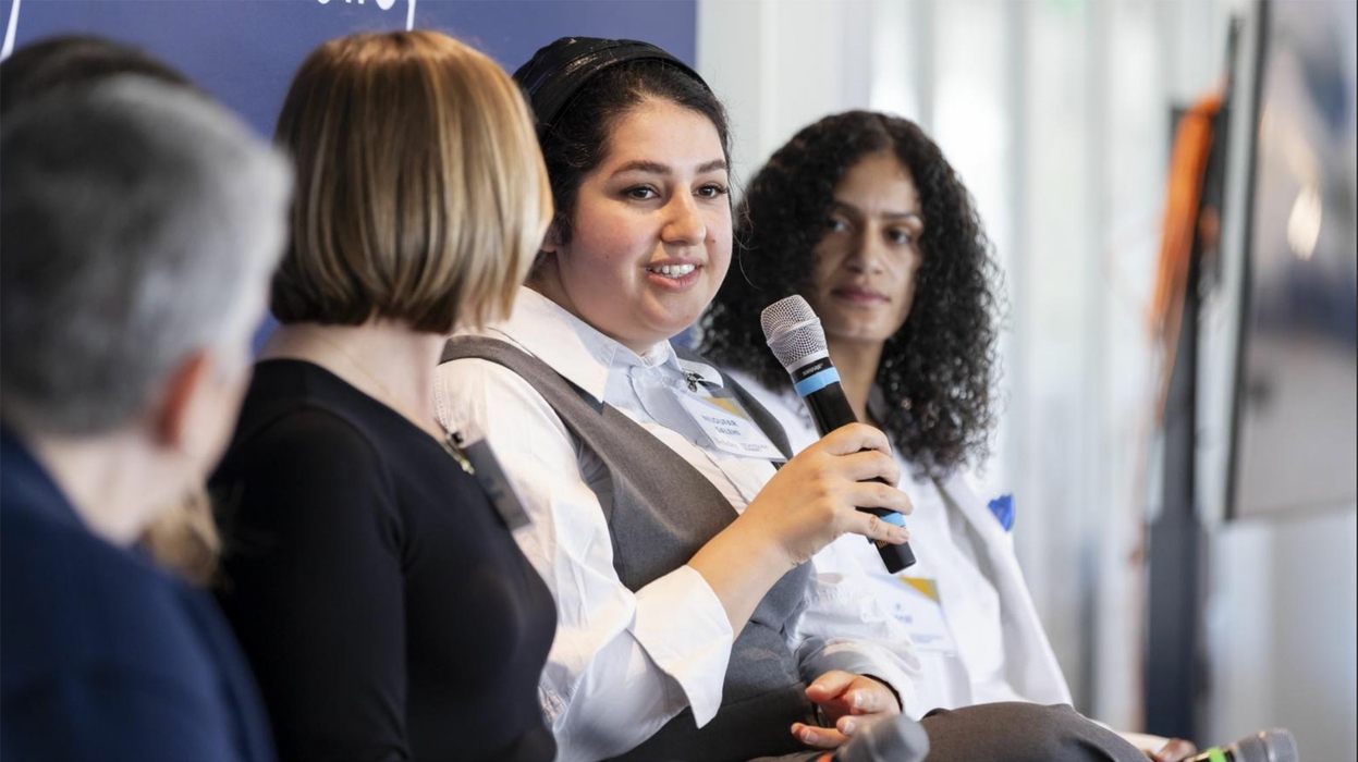Woman holding microphone in panel setting with other panelist looking in her direction.