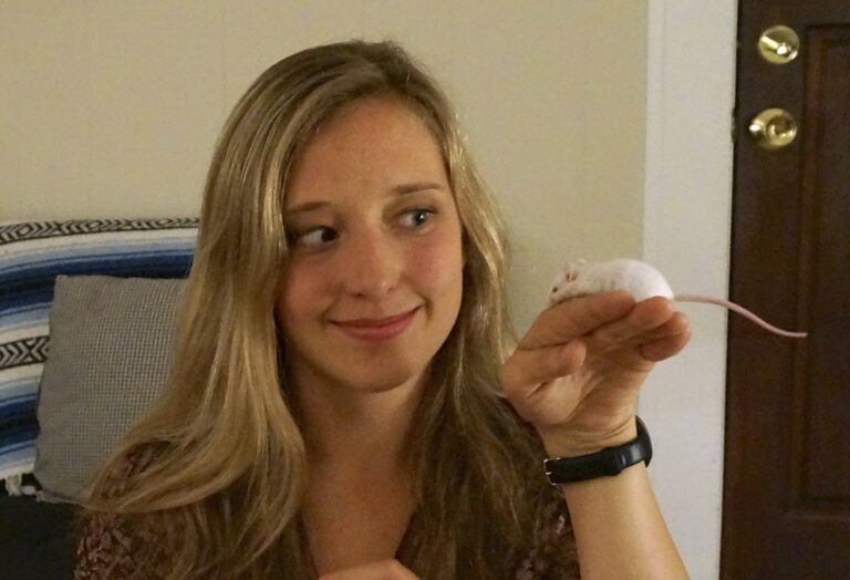 blond woman facing screen but looking to side at a white mouse on her hand