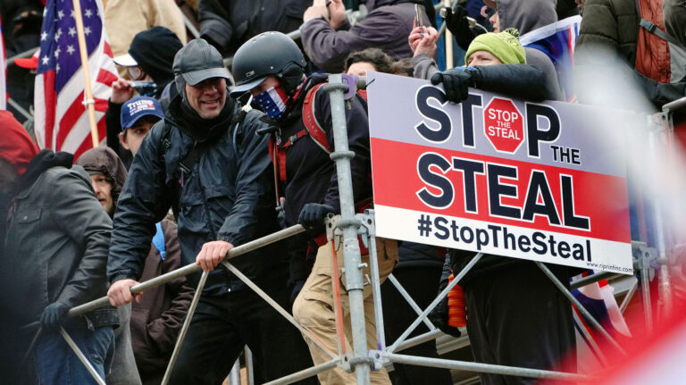 A mob of men, some wearing assault gear, pass by a "Stop the Steal" placard as they attack the US Capitol.