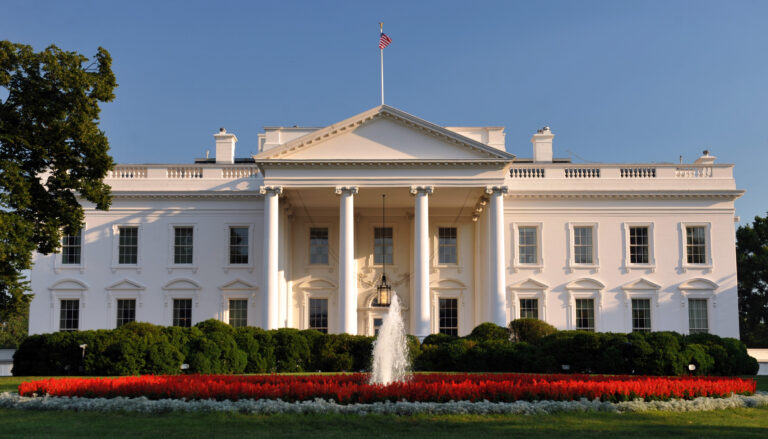 The north entrace to the White House, in late-day sunlight, with a garden and fountain in the foreground and a deep blue sky in the background