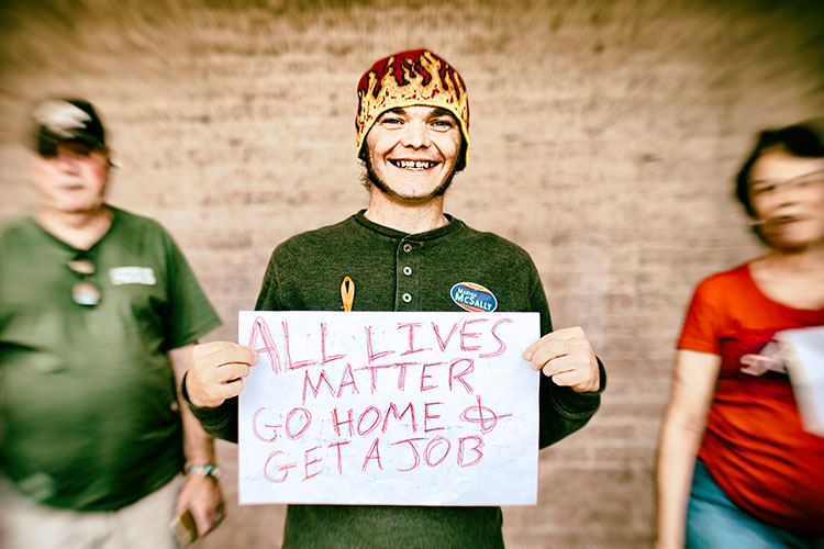 young man in hat holds hand-made sign that says: "All Lives Matter | Go Home & Get a Job"