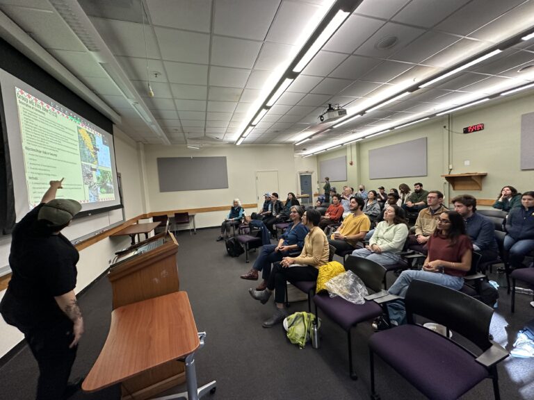 A student points to a powerpoint slide while giving a talk to a crowd of listeners.