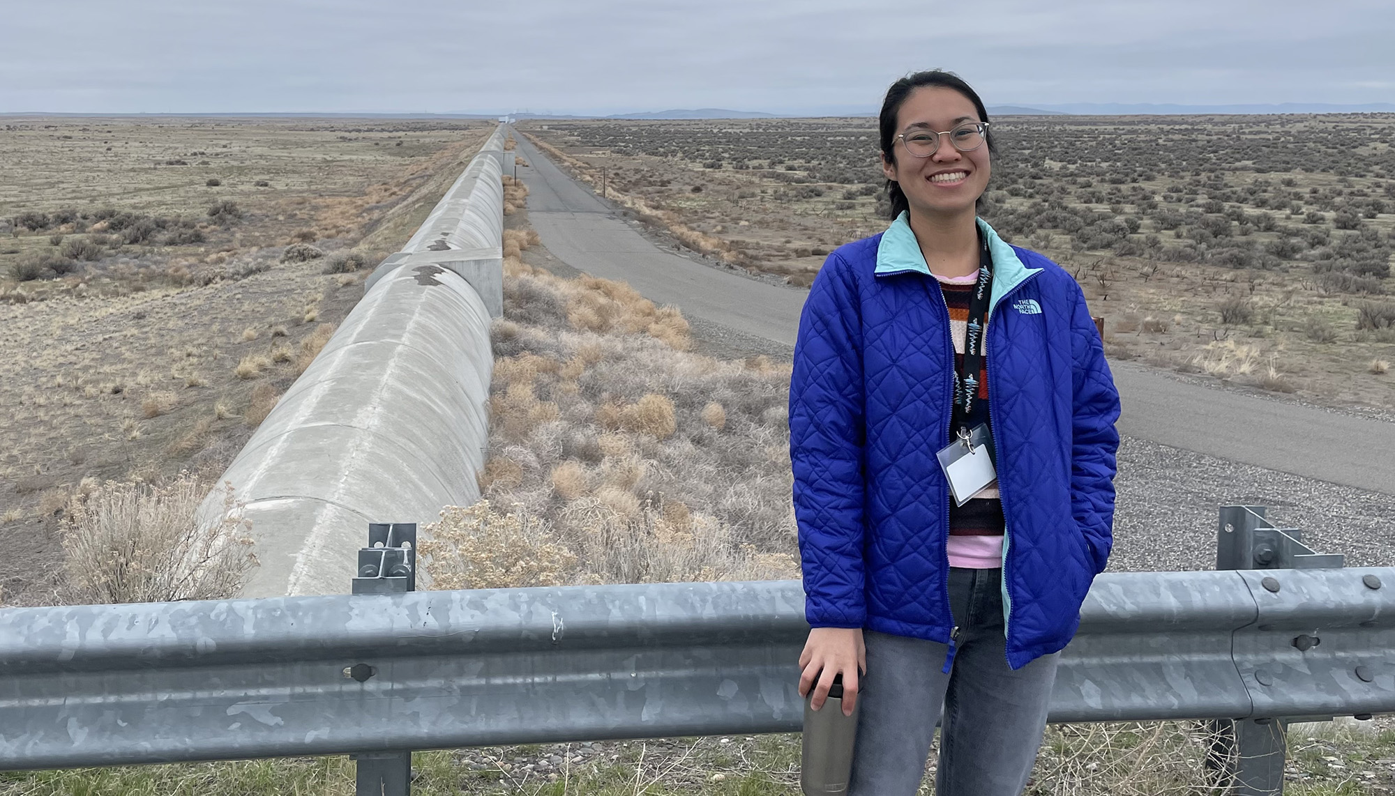 a woman in blue jacket standing in a treeless plain with a concrete conduit in the background