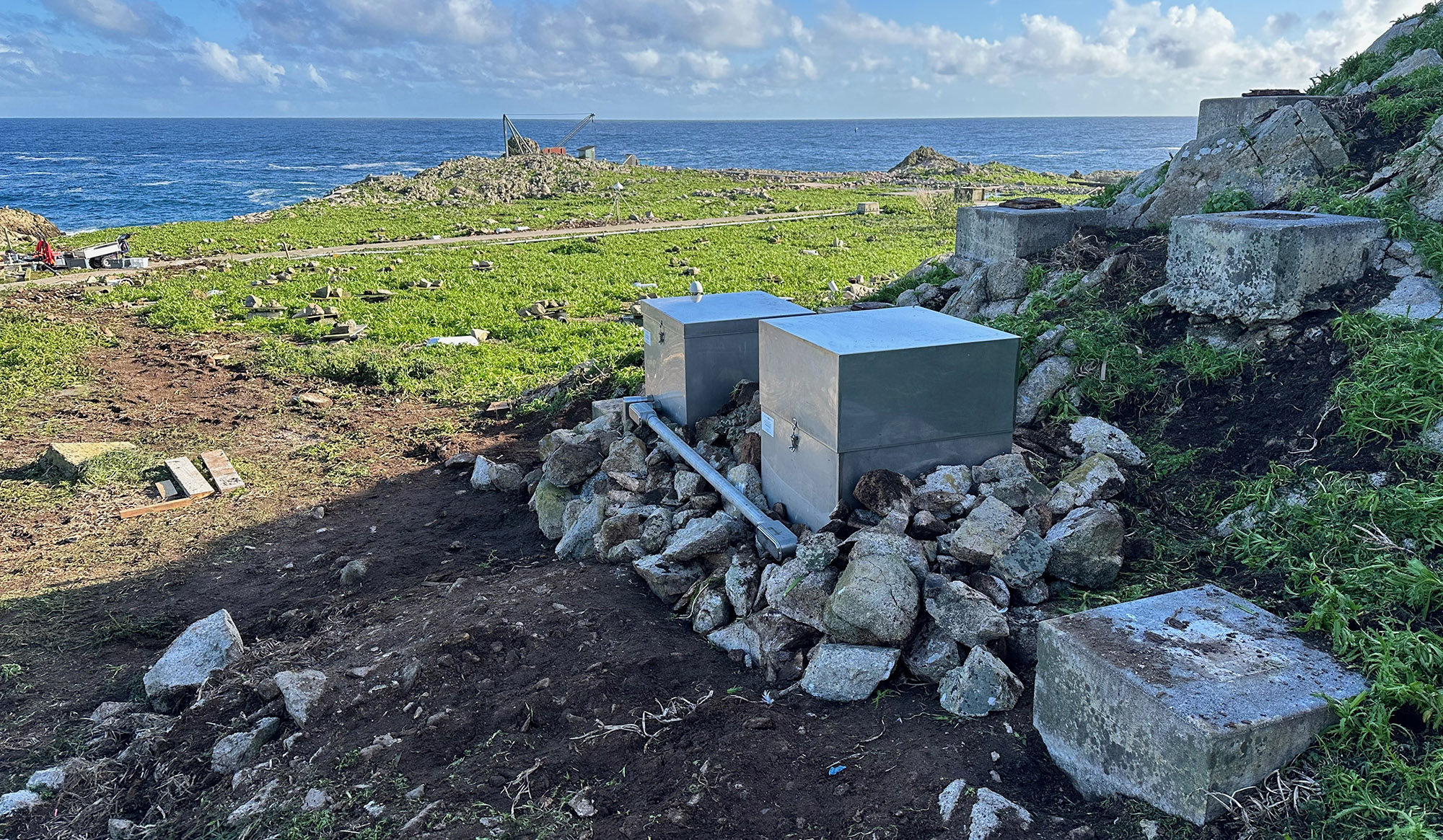 2 stainless steel boxes sit among rocks on a hillside overlooking the ocean