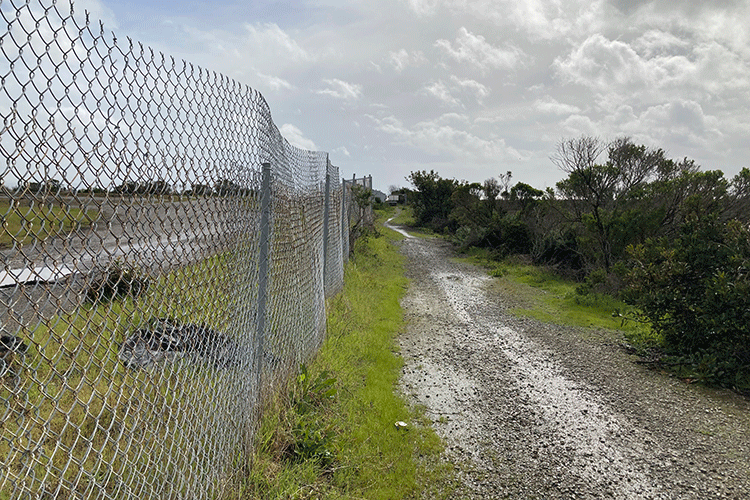 A photo shows a chain link fence that runs alongside a dirt road. On the other side of the fence from the road is an open field that is partially covered in grass but with some patches of a gravel-like substance showing through.