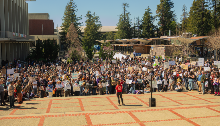 a person in a red shirt addresses a crowd of people holding placards in Sproul Plaza