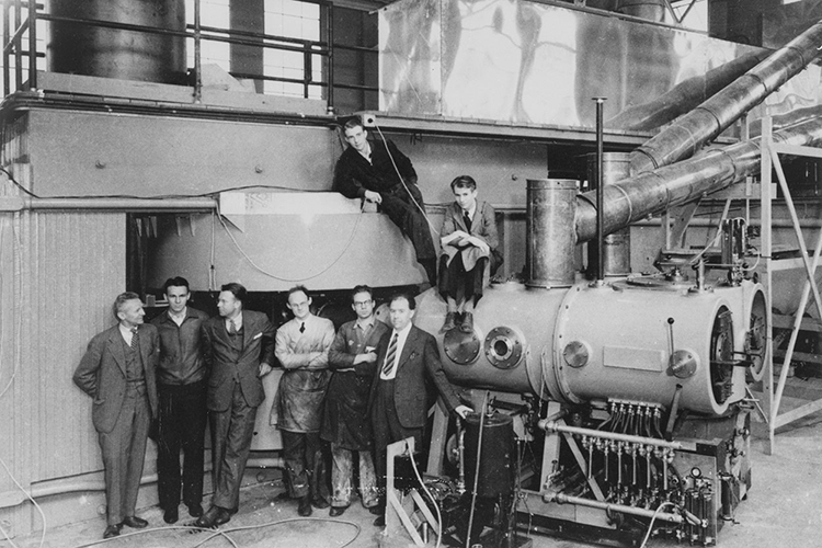 8 people standing in a line next to the 60-inch cyclotron, Ernest Lawrence being the third person from the left 