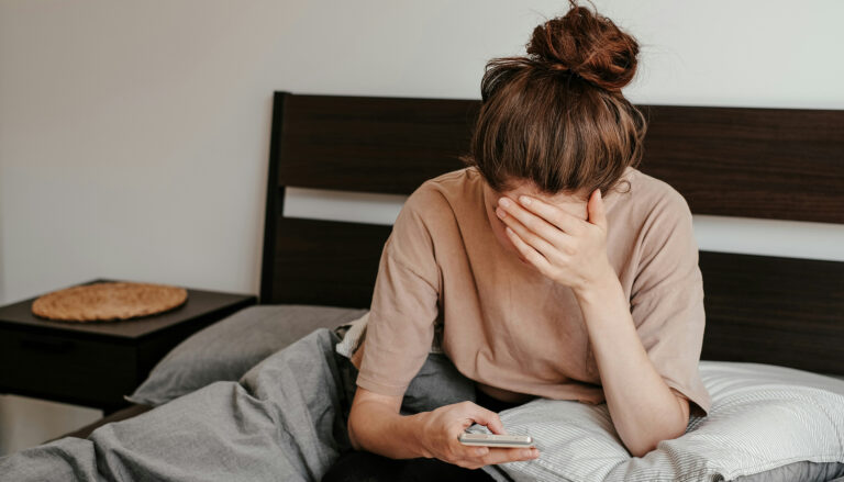 a woman sits in bed looking down at her phone with a hand covering her eyes