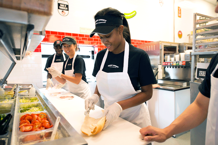 fast food worker, smiling, stands at a counter making a sandwich. Colleagues are working to her side.