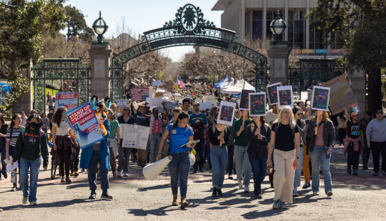 a crowd carrying signs marches though Sather Gate