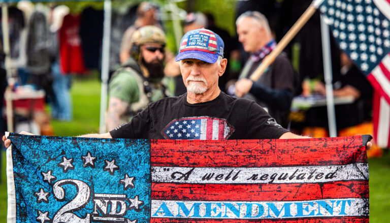 A bearded man in a baseball cap holds a U.S. flag that bears the words "A well regulated 2nd Amendment"
