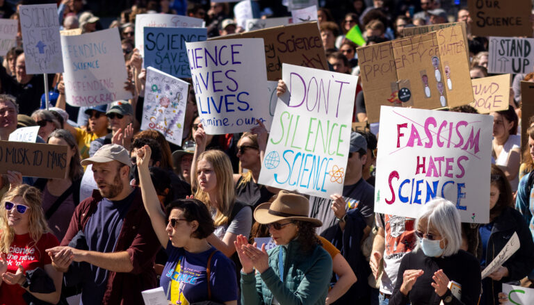 protesters in a crowd with signs reading Science Saves Lives, Don't Silence Science and Fascism Hates Science