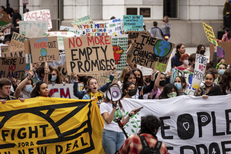 A throng of students protest, with one student holding a bullhorn. They carry a variety of signs, but the prominent one says: "No more empty promises."