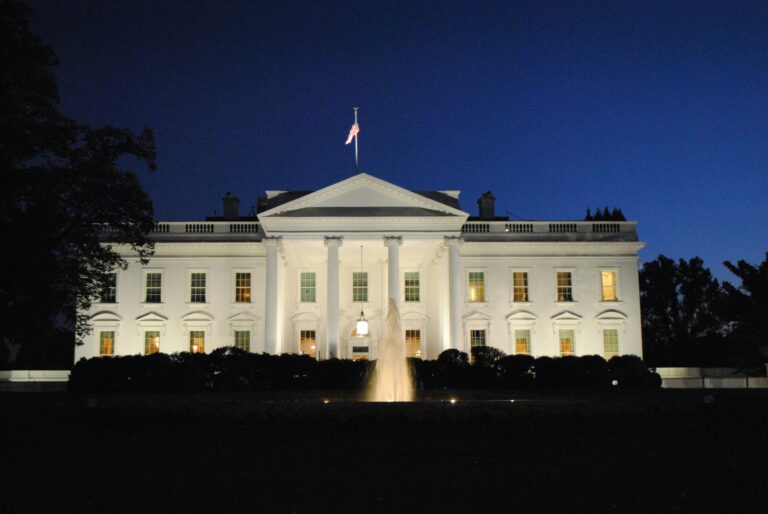 The White House in Washington D.C. at night, illuminated with lights and with a fountain in the foreground shooting water into the air.