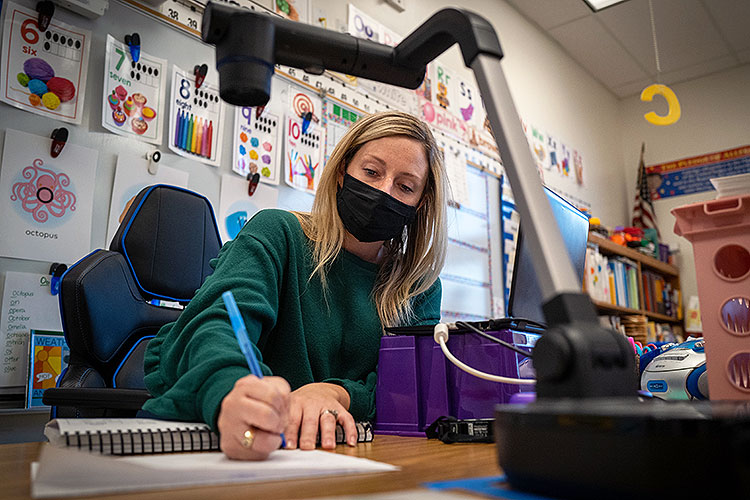 masked teacher working at her desk, surrounded by technology and colorful learning posters