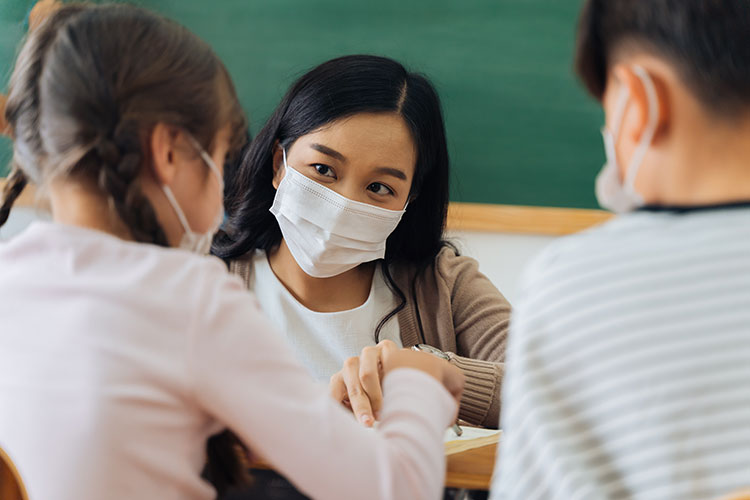 a teacher and two young students, all wearing masks to protect against COVID-19 infection
