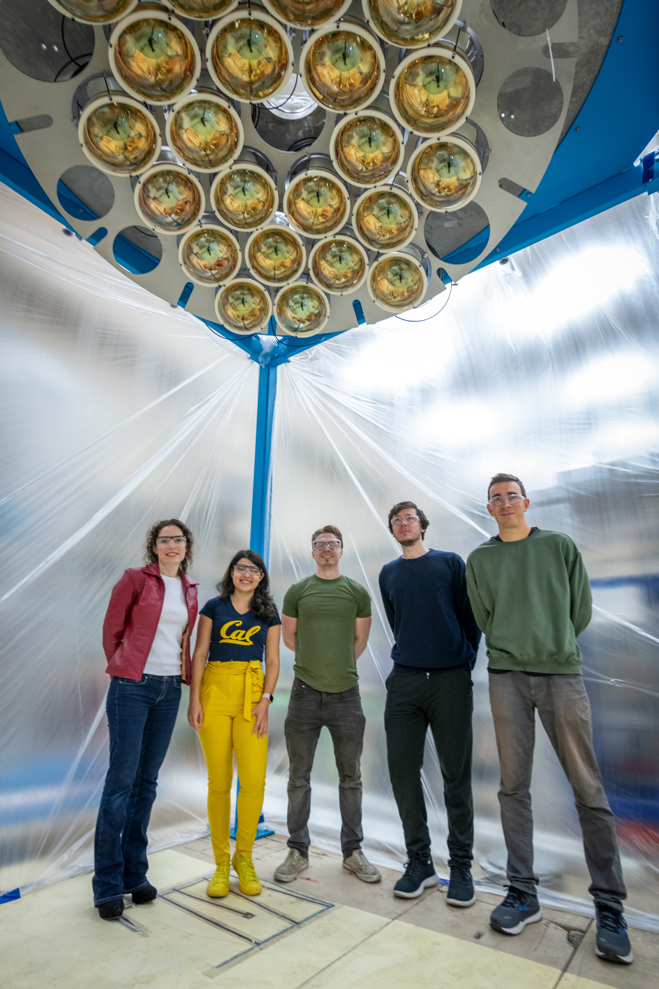 5 people wearing goggles inside a plastic tent below a shiny bank of amber photomultiplier tubes