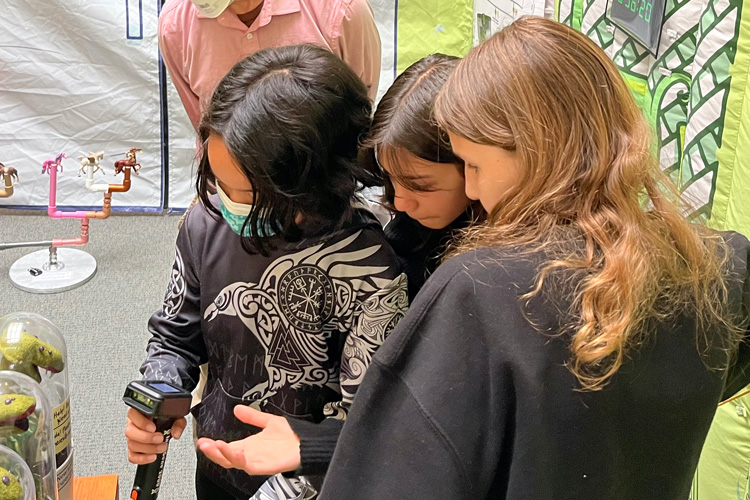 a dark-haired boy in a mask with two girls looking at a digital measuring device