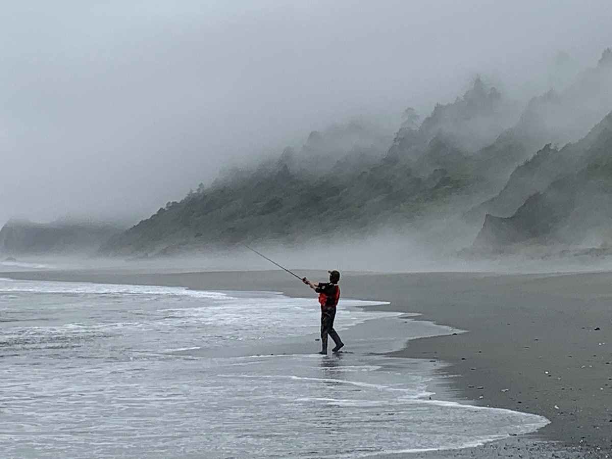 Person standing at the water's edge with fishing pole extended into the water; mist along the shore.