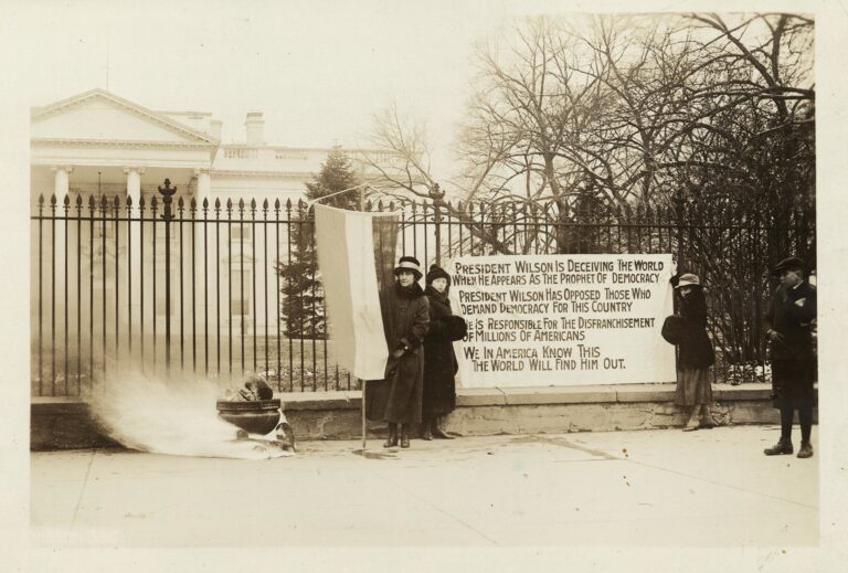 Three women demonstrate in front of the White House fence in a sepia photo. A watchfire burns, one woman holds a flag, and two others hold up a banner that reads "President Wilson is deceiving the world when he appears as the prophet of democracy. President Wilson has opposed those who demand democracy for this country. He is responsible for the disfranchisement of millions of Americans. We in America know this. The world will find him out."