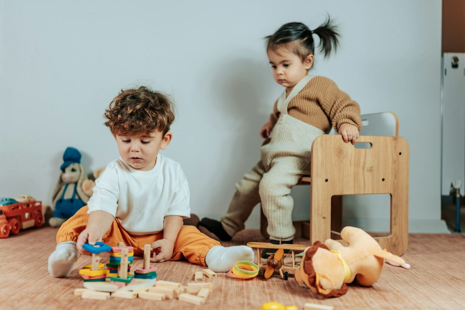 two toddlers in a room, with one looking over the shoulder of the other as they play with blocks. the child playing with the blocks has amassed several in front of him.