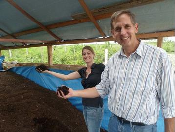 Kramer and Andersen stand in a covered area filled with compost and show handfuls of it to the camera