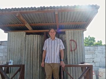 Andersen wears a striped shirt and stands under the overhang of a sheet metal latrine building