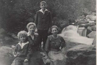 2 women and a child sitting, and 1 child standing on rocks in front of a creek.