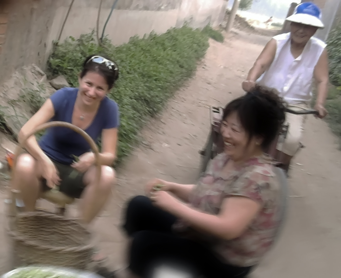 A woman speaks to one woman putting beans into a basket and one woman riding a bicycle.