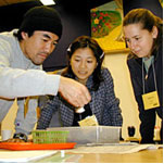 3 people standing over table looking at something on the table