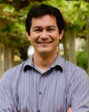 Prof. Tsutsui is wearing a blue shirt with thin white stripes, smiling in front of a background of trees