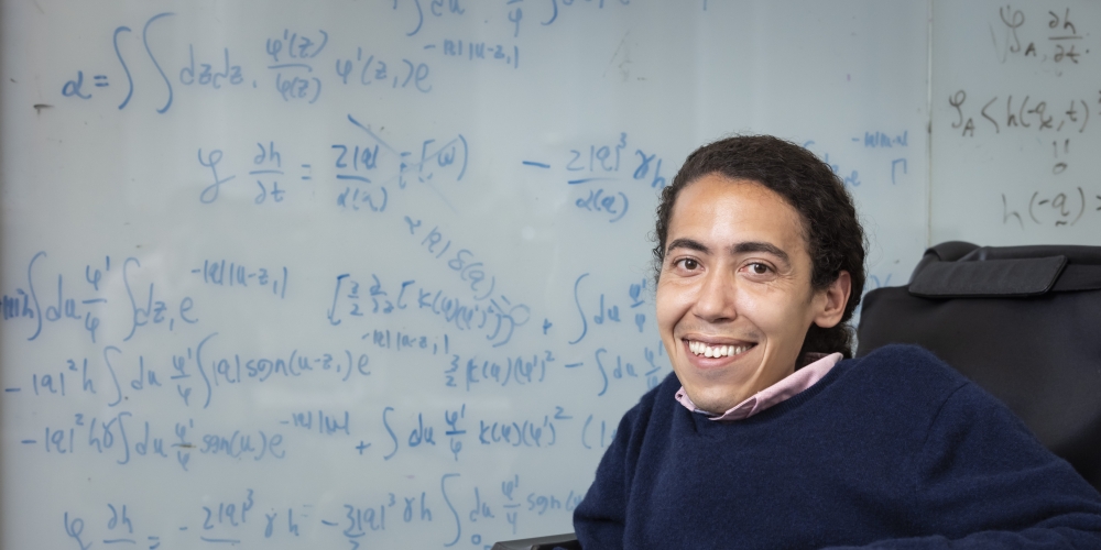 Professor Ahmad Omar sitting in front of a glass whiteboard marked with equations related to his research. 
