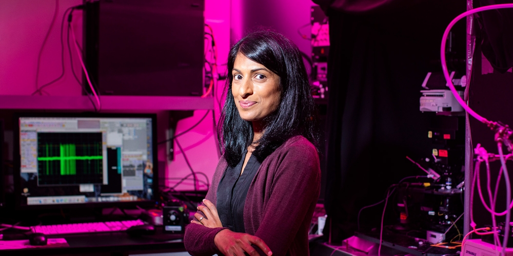 Teresa Puthussery in lab setting with pink filtered lighting and machinery in background.