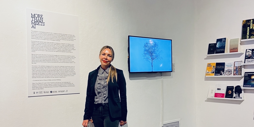 Woman with blond hair standing in an art gallery next to books, a video monitor, and wall text.
