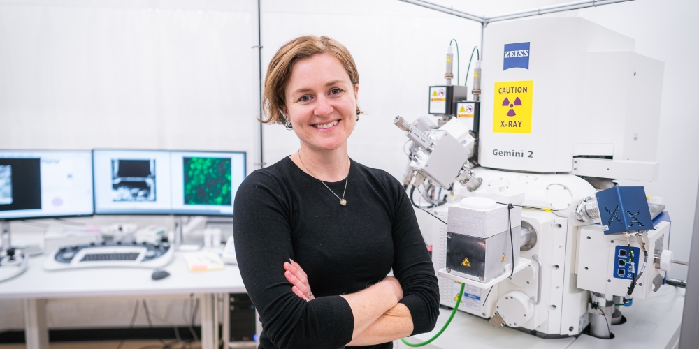 Assistant Professor Bronwyn Lucas stands with arms crossed next to a Zeiss Crossbeam cryo-FIBSEM.