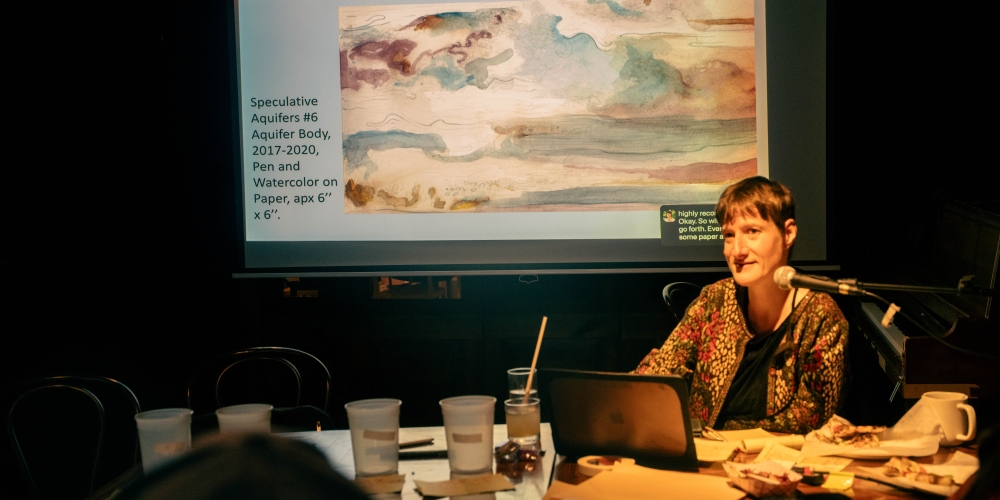 Image of a white disabled woman in her forties. She is speaking into a microphone giving a presentation. Image is of Sunaura Taylor.